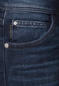 Tmavě modré džíny z denimové látky s mírně opotřebovanou texturou. Mají stříbrné příslušenství a černou etiketu s nápisem "Calvin Klein Jeans."