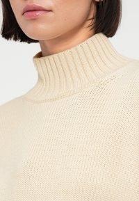 Μπεζ πλεκτό πουλόβερ με ψηλό ριμπ κολάρο. Η υφή είναι λεία με λεπτό, ομοιόμορφο σχέδιο ραφής. Χωρίς εμφανείς διακοσμήσεις ή μεταλλικά μέρη.
