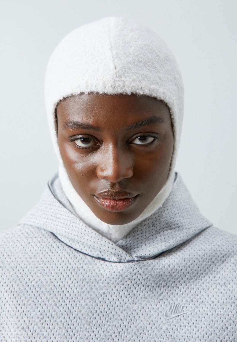 Nahaufnahme einer Person mit dunkler Haut, die eine weiße strukturierte Sturmhaube und einen hellgrauen Nike-Hoodie trägt und direkt nach vorne schaut.