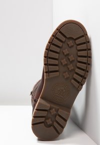 La semelle en caoutchouc marron présente un design texturé avec des rainures profondes et un logo circulaire. La semelle démontre une construction robuste et durable.