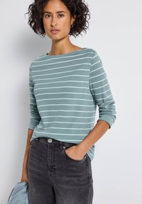 Hellblaues Langarmshirt mit weißen horizontalen Streifen, Rundhalsausschnitt und lässiger Passform. Trägt dazu schwarze Jeans mit Vordertaschen.