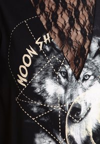 Svart plagg med en vargtryck och texten "MOON SH". Spetsdetaljer vid halsringningen ger textur och visuell kontrast med en geometrisk kontur.