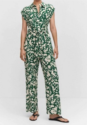 Person iført en grøn og hvid abstrakt mønstret ærmeløs jumpsuit med bindebælte og sorte sandaler.