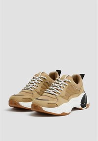 Chaussures de sport couleur beige avec des lacets blancs, présentant une tige texturée, des accents noirs contrastants et une semelle épaisse blanche et brune.