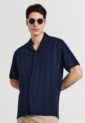 Homme portant une chemise à manches courtes bleu marine et un pantalon beige, posant avec des lunettes de soleil et les mains derrière le dos.
