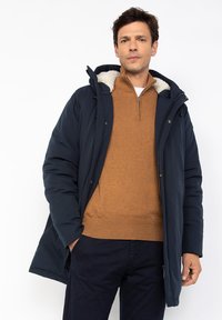 Hombre con parka azul marino con capucha de piel sintética, suéter marrón de medio cremallera y pantalones oscuros, de pie contra un fondo blanco.