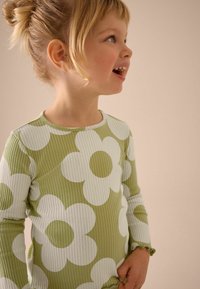 Grünes, geripptes Langarmshirt mit großen weißen Blumenmustern, mit Rüschensäumen und rundem Ausschnitt, getragen von einem Kind.