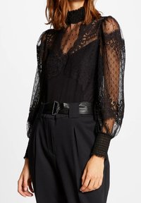 Blusa nera trasparente con pizzo stampato, collo alto e maniche a sbuffo. Presenta polsini arricciati e un corpo in tessuto liscio, abbinata a una cintura nera.