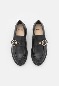 Mocasines de cuero negro con punta redonda, detalle de herrajes dorados y suela texturizada. Presentan una correa en el empeine para el cierre.