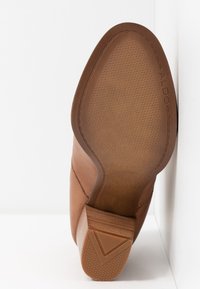 Semelle de chaussure en cuir marron avec grip texturé, talon carré en bois et nom de la marque "ALDO" embossé près du bord.