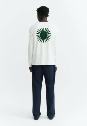 Uomo di spalle che indossa una camicia bianca a maniche lunghe con un cerchio verde a raggi di sole sulla schiena e pantaloni blu scuro.