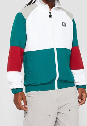 Man draagt een colorblock jas met witte, teal, rode en grijze stukken, dicht met een rits, gecombineerd met lichtbeige broek.