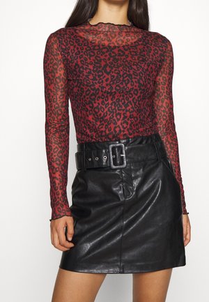 Vrouw draagt een rood-zwarte doorschijnende top met luipaardprint en lange mouwen, ingestopt in een zwarte leren minirok met een brede riem en een vierkante gesp.