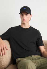 Black short-sleeve t-shirt with a round neckline, paired with olive green pants. A black cap with a blue logo sits atop. Textured backdrop.
