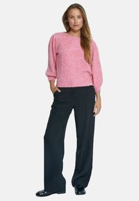 Rosa, strukturiertes Strickpullover mit langen, puffierten Ärmeln und rundem Ausschnitt, kombiniert mit schwarzen, weit geschnittenen Hosen und glänzenden schwarzen Schuhen.