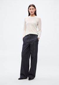 Bluză bej deschis cu mâneci lungi, asortată cu pantaloni din denim închiși la culoare, cu croială largă. Pantalonii au un material texturat și o curea neagră contrastantă.