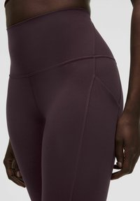 Nahaufnahme einer Person, die hoch geschnittene, dunkelviolette Leggings mit sichtbaren Nähten und einer Seitentasche trägt, vor einem schlichten Hintergrund.