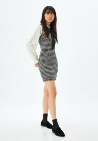 Sort og hvidt houndstooth-kjole med en tætsiddende silhuet, frynset kant og en sløjfe-accent. Langærmet hvid skjorte synlig under.