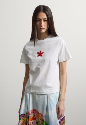 Frau mit langen dunklen Haaren trägt ein weißes T-Shirt mit rotem Seestern-Detail und einen plissierten Rock mit buntem Küstenstadt-Print.