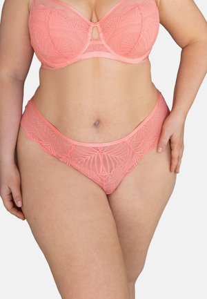Woman wearing matching coral lace bra and high-cut panties, standing with one hand on hip against white background.