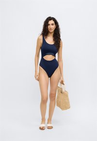 Μπλε σκούρο ολόσωμο μαγιό με λεπτομέρεια κοψίματος και λωρίδα με λογότυπο. Το μοντέλο κρατά πλεκτή τσάντα και φορά ανοιχτόχρωμα σανδάλια.