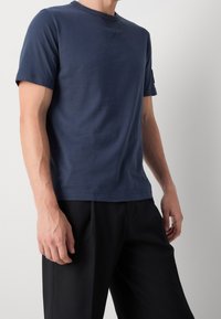 Mies, joka pukeutuu tummansiniseen lyhythihaiseen paitaan ja mustiin housuihin, seisoo tasaisella vaalealla taustalla.