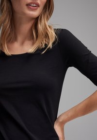 Woman with shoulder-length blonde hair wearing a fitted black long-sleeve shirt with her hand resting on her hip against a gray background.