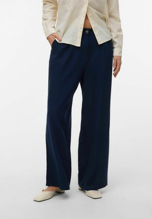 Osoba odjevena u široke teget hlače širokih nogavica, bež košulju s gumbima i bež cipele s kvadratnim prstima, stoji s jednom rukom u džepu.