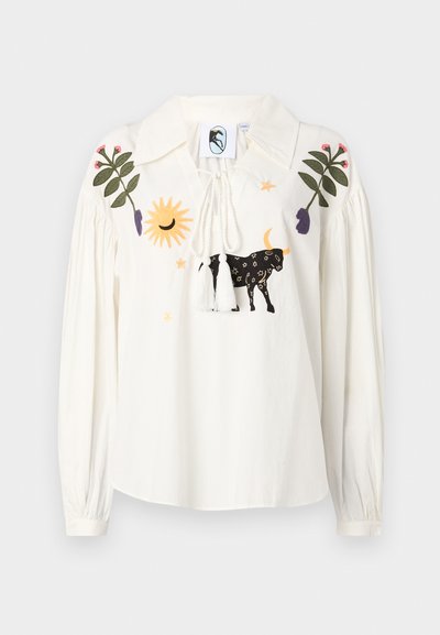 Blusa bianca a maniche lunghe con colletto a punta, caratterizzata da motivi ricamati multicolore tra cui una mucca, un sole e foglie. Chiusura con coulisse e frange.