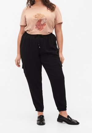 Femme portant un t-shirt beige avec un art abstrait de visage, un pantalon cargo noir avec taille à cordon de serrage, et des mocassins noirs sans lacets, debout sur fond blanc.