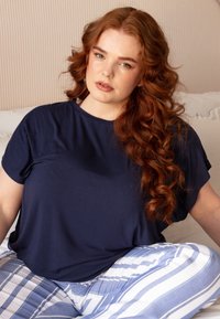 Marineblauw T-shirt met korte mouwen van zacht materiaal, gecombineerd met wit en blauw gestreepte broek. Model zit tegen een lichte achtergrond.
