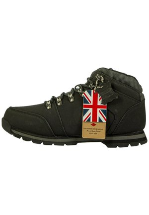 Czarne buty trekkingowe z teksturowanym materiałem, wyraźnym przeszyciem i metalowymi oczkami. Posiadają metkę z flagą Zjednoczonego Królestwa oraz solidną gumową podeszwę.