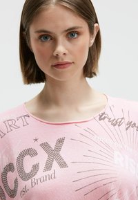 Junge Frau mit kurzen braunen Haaren und blauen Augen, die ein pinkes Shirt mit Text und grafischen Designs auf einem hellen Hintergrund trägt.