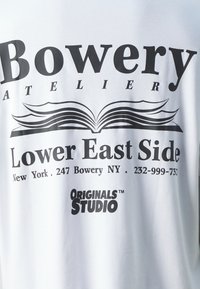 Balta medvilninė marškinėliai su drąsiais juodais užrašais. Dizainas apima atidarytą knygą ir pavadinimus "Bowery", "Lower East Side" ir "ORIGINALS STUDIO."