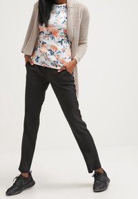 Kobieta w czarnych spodniach, czarnych trampkach, kwiecistej bluzce i beżowym swetrze z dzianiny, stoi z rękami w kieszeniach na tle białym.