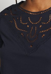 Námořnická modrá tričko s náročnými výřezy vyšívanými kolem výstřihu, s texturovaným povrchem a podrobnými dekorativními vzory.