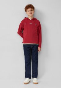 Rode hoodie met een witte binnenlaag, vooraan een logo en zijaccenten, gecombineerd met donkerblauwe jeans en witte sneakers met beige zolen.