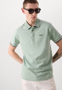 Světlezelené polo tričko s texturovaným límcem, černobílými pruhovanými akcenty a malým logem "BOSS" na hrudi. Krátké rukávy a hladká tkanina.