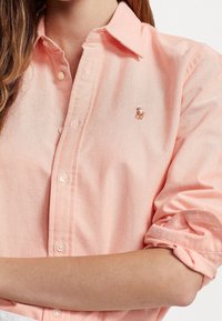 Světle růžová košile na knoflíky vyrobená z jemné tkaniny s klasickým límečkem. Má ohrnuté rukávy a vyšívané logo na hrudi.