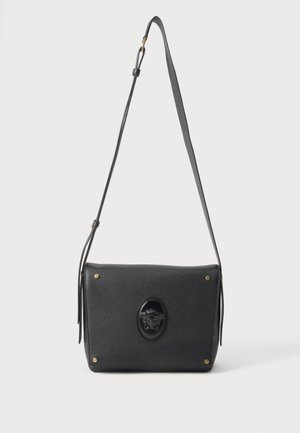 Sac à épaule en cuir noir avec bandoulière ajustable, surface texturée, clous dorés aux coins et emblème central embossé en forme de tête de Méduse.