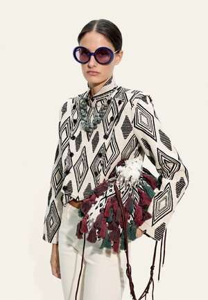 Kobieta w czarno-białej kurtce z geometrycznym wzorem, białych spodniach, dużych okrągłych fioletowych okularach przeciwsłonecznych, masywnym naszyjniku z kamienia, trzymająca torebkę ozdobioną frędzlami.