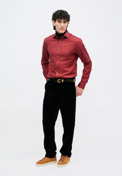 Crvena košulja na dugmad, crne baršunaste hlače, crni remen sa zlatnim hardverom i smešane tenisice. Model stoji na neutralnoj pozadini.