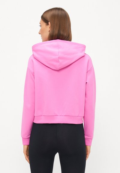 Skrajšani kapuzen pulover v svetlo rožnati tkanini z gladko teksturo. Ima kapuco in prilagojene manšete, kombiniran z črnimi legicami.