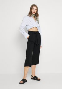 Ljusblå kortärmad skjorta med knuten front, svarta vida byxor med knuten bälte och svarta sandaler med tjock sula.