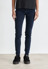 Dark blue slim-fit jeans made of denim fabric, featuring a smooth texture and minimal detailing, paired with black sneakers.