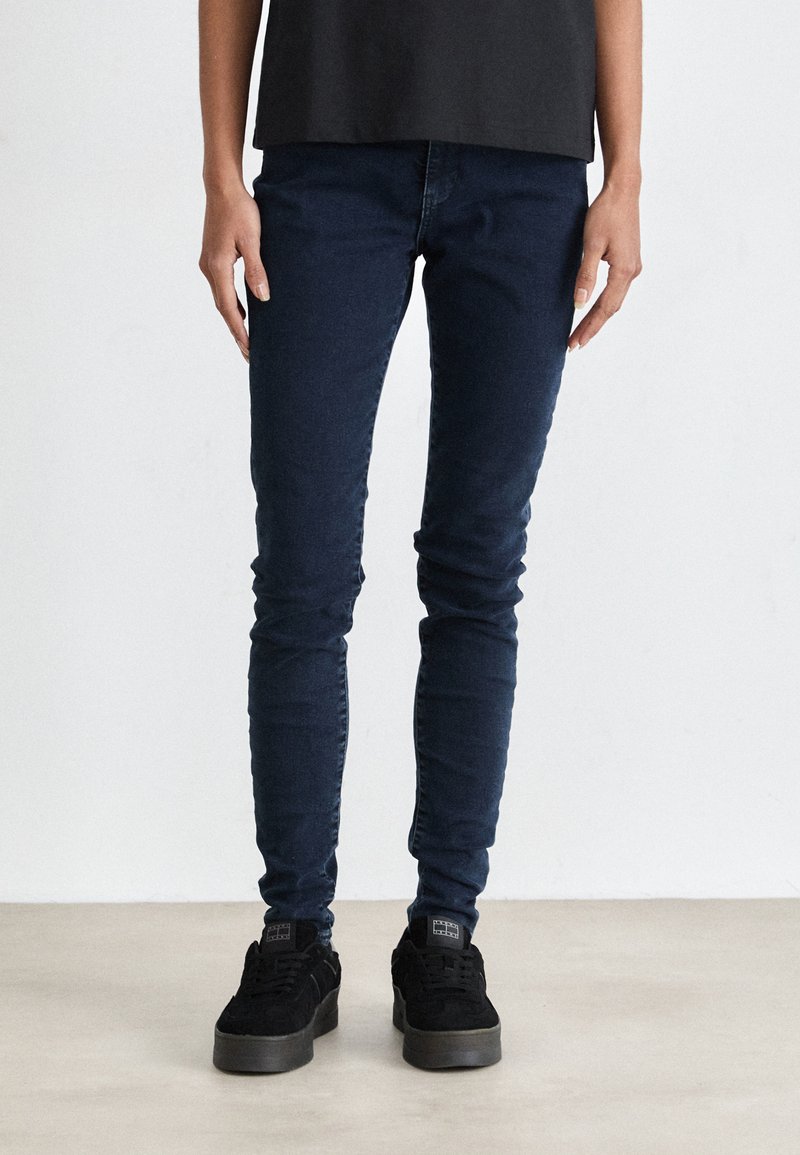 Dark blue slim-fit jeans made of denim fabric, featuring a smooth texture and minimal detailing, paired with black sneakers.