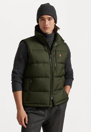 Olive green puffer vest with high collar, front zip pockets, and logo detail. Worn over a dark gray turtleneck and paired with gray trousers.