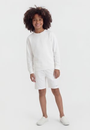 Uśmiechnięte dziecko o kręconych włosach stoi, ubrane w białą bluzę, białe szorty z logo Nike oraz białe buty wsuwane, na jednolitym tle.
