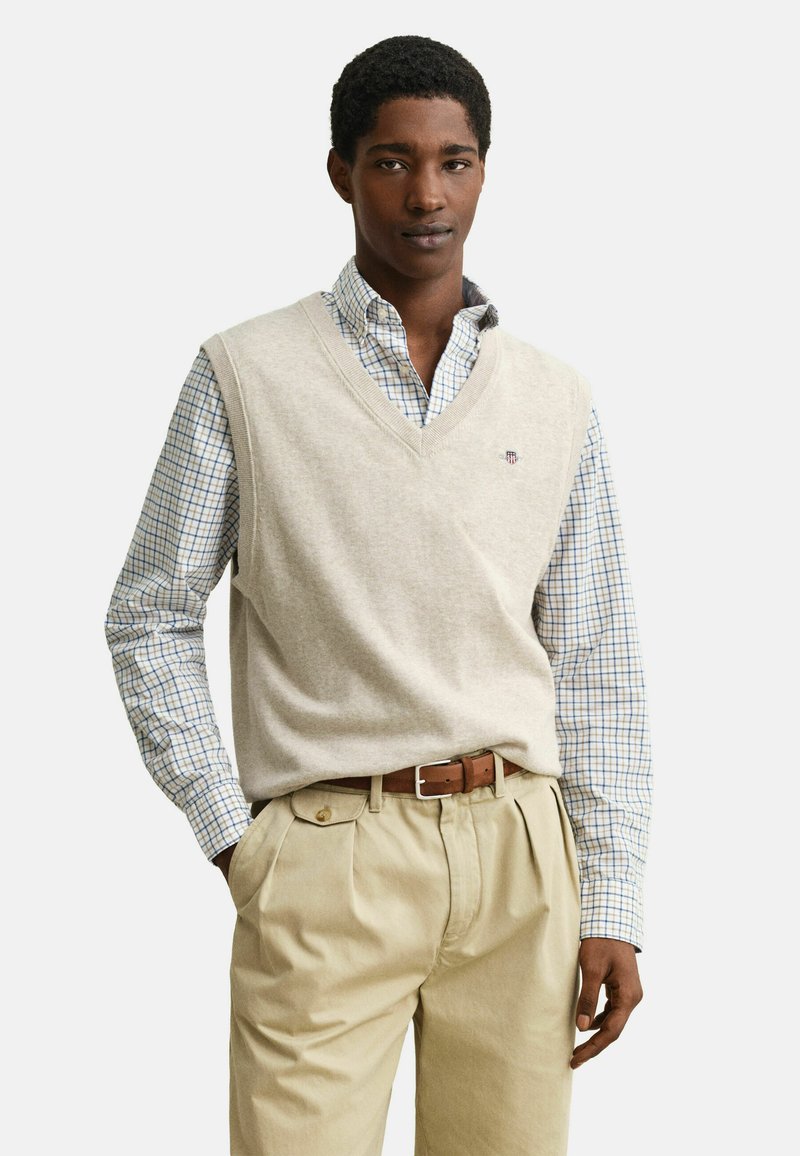 Jeune homme portant un gilet en tricot beige à col en V par-dessus une chemise blanche à carreaux bleus, un pantalon beige plissé et une ceinture marron, debout avec la main dans la poche.