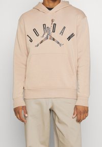 Sudadera con capucha beige con bolsillo tipo canguro, que presenta un gran logo negro "JORDAN" y un gráfico abstracto de un jugador de baloncesto en el centro.